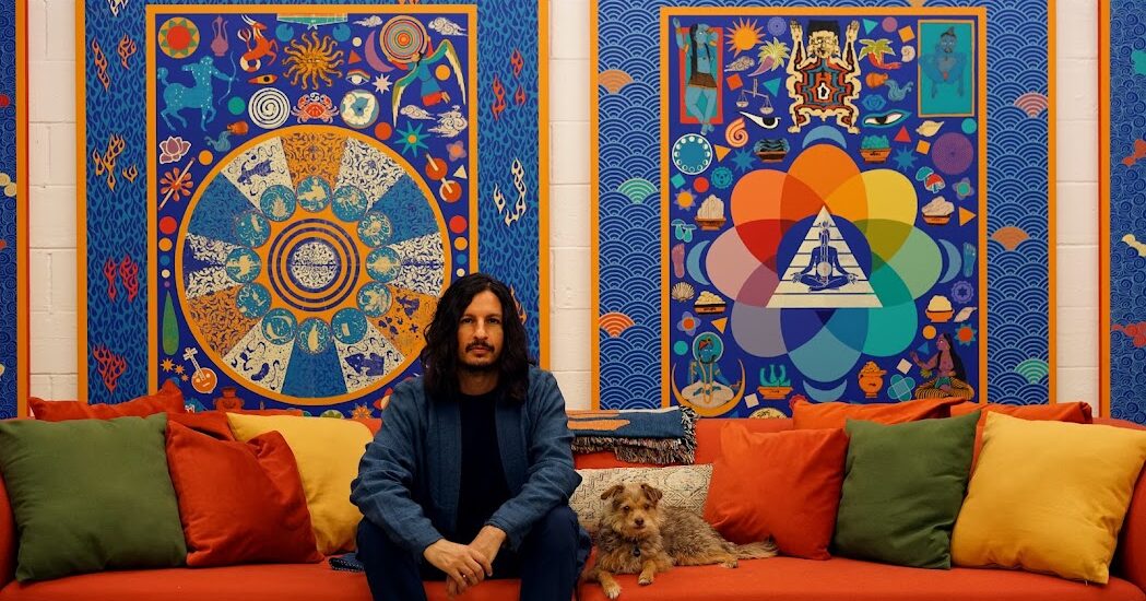 a Mann sitting on a couch with a dog in from of paintings he created.