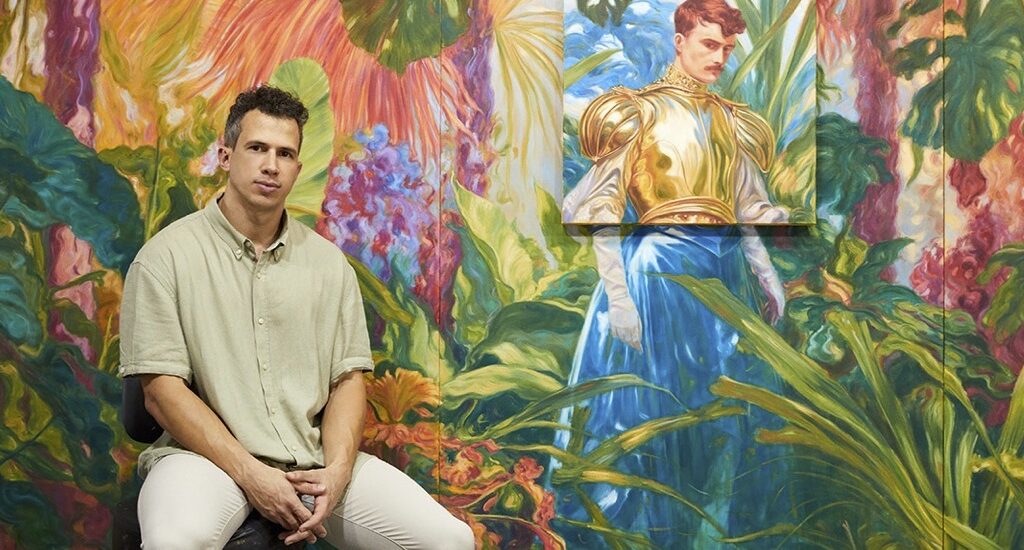 Marlon Portales sitting in front of a luswh painting of landscapes and figurtatism